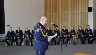 Sala konferencyjna. Mężczyzna w średnim wieku, stoi przy mikrofonie, w rękach trzyma kartkę. Ubrany jest w granatowy mundur Straży Miejskiej, na nosie ma okulary. Za nim siedzi dziesięć osób na krzesłach. Wszyscy ubrani są w granatowe mundury Straży Miejskiej.