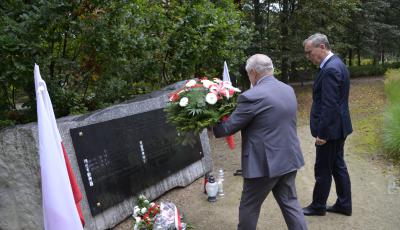 Park. Przed pomnikiem ofiar katyńskich stoi dwóch mężczyzn w średnim wieku. Jeden z nich trzyma wiązankę z białymi kwiatami. Przy pomniku stoją znicze i leżą biało-czerwone kwiaty. Po obu stronach pomnika stoją na stojaku biało-czerwone flagi.