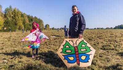 Łąki Oborskie. Chłopiec i dziewczynka w wieku szkolnym stoją obok siebie. Dziewczynka trzyma w rękach różowy latawiec w kształcie trójkąta. Chłopiec trzyma w ręku latawiec w kształcie sześciokąta , na którym namalowany jest kolorowy motyl. W tle drzewa i błękitne niebo.