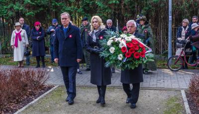 Park. Dwóch mężczyzn i jedna kobieta stoją obok siebie w rzędzie. Jeden z mężczyzn w rękach trzyma duży wieniec z biało-czerwonymi kwiatami.