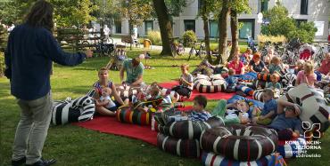 Grupa dzieci siedzi na wielkich poduchach rozrzuconych na trawie w Parku Zdrojowym. Przed nimi stoi mężczyzna w średnim wieku i coś opowiada.