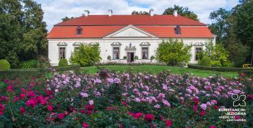 Widok na niski dwór z pomarańczowym dwuspadowym dachem. Przed budynkiem znajduje się duża rabata z fioletowych kwiatów.