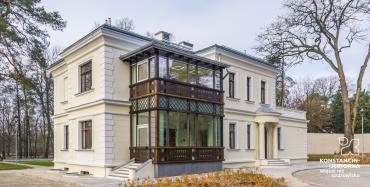 Widok na boczną elewację budynku willi Kamilin z ozdobną drewnianą werandą. W tle rosnące wysokie drzewa
