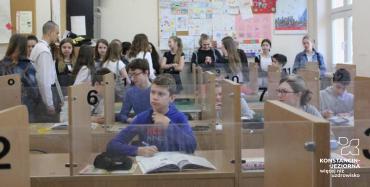 Szkolna sala lekcyjna z wydzielonymi stanowiskami poprzedzielanymi przezroczystymi płytami, na sali dużo dzieci