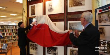 Dwaj mężczyźni zdejmują flagę Polską ze ścianki, na której wiszą fotografi wraz z opisami, w tle widoczne biblioteczne regałty z książkami
