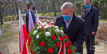 Mężczyzna składa wieniec, za nim widoczny inny stojący, obok flagi polska w stojaku