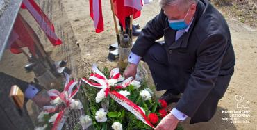 Mężczyzna w przysiadzie składa wieniec, przed pomnikiem z tablicą, obok flagi polskie w stojaku