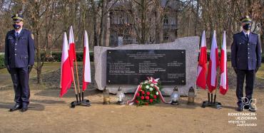 Pośrodku poziomy pomnik kamienny z czarną tablica z tekstem, po bokach flagi polski w stojakach oraz dwaj mężczyźni w mundurach straży miejskiej