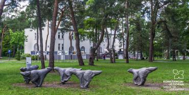 Trzy duże, metalowe rzeźby, które mają postać biegnących wilków. Rzeźby ustawione są na trawie, wśród wysokich drzew. W tle widać wille Hugonówkę