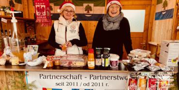 Stoisko handlowe udekorowane świątecznie, przy ladzie stoją dwie kobiety w ciepłych strojach i z nakryciami głowy, na dole lady baner z napisamei w języlku polskim i niemieckim