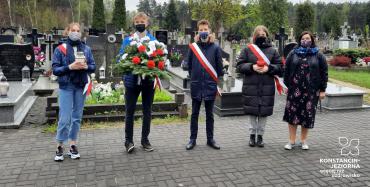 Pięc osób stojących na cmentarzu obok siebie. Dwie osoby trzymają znicz, jedna biało-czerwoną wiązankę z kwiatów . W tle las.