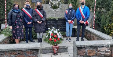 pięć osób stojacych obok pominka ubranych w kurtki oraz biało-czerwone szarfy. Na pomniku leży wiązanka patriotyczna