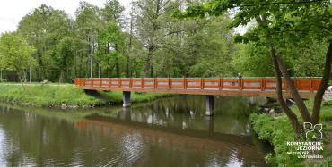 Drewniany, duży  mostk w parku, pod nim płynie rzeka, po obydwu stronach brzegu liczne drzewa, zdjęcie ilustrujące opisaną w artykule sytuację