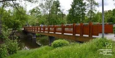 Drewniany mostek w parku, pod nim rzeka, na brzegach drzewa i krzewy, zdjęcie ilustrujące opisaną w artykule sytuację