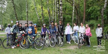 Wycieczka rowerowa klkunastu uczniów. Dzieci stoją przy swoich rowerach. W tle rosną wysokie drzewa.