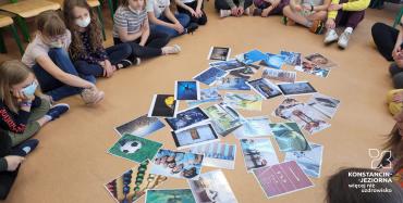 Dzieci siedzące w okręgu na podłodze sali lekcyjnej, spoglądają na rozsypane wewnątrz kolorowe fotografie.