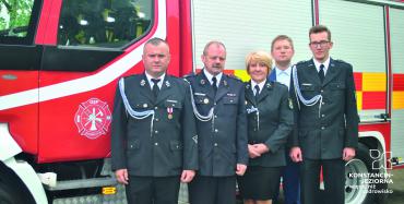 Pięć osób (czterech mężczyzn i jedna kobieta) stoi w mundurach strażackich na tle wozu strażackiego.
