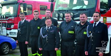 Siedmiu mężczyzn stoi w mundurach strażackich na tle wozów strażackich.