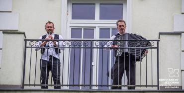 Na balkonie dwóch muzyków, na lewo grający na klarnecie, na prawo grający na akordeonie, za nimi duże przeszklone drzwi.
