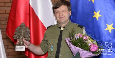Uśmiechnięty mężczyzna w mundurze harcerskim w jednej ręce trzyma statuetkę w kształcie dębu, a w drugiej – kwiaty, teczkę z dyplomem i torbę podarunkową.