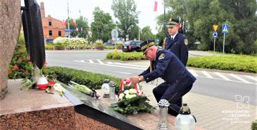 Dwóch mężczyzn, ubranych w stroje mundurowe Straży Miejskiej składają wieniec przed pomnikiem. 