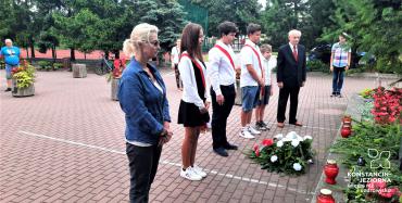 Siedem osób, w tym czterech uczniów stoi na baczność przed pomnikiem. Przed nimi leży wiązanka z biało-czerwonych kwiatów.
