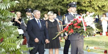 Mężczyzna, ubrany w wojskowy strój mundurowy maszeruje niosąc w rękach wieniec z biało-czerwonych kwiatów. Za nim podążają inni ludzie. 