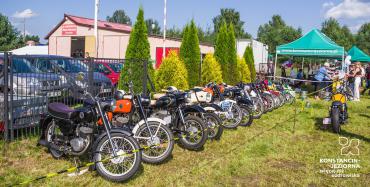 Dwanaście motocykli różnego koloru stoi w rzędzie na trawie przy metalowym ogrodzeniu. Odgrodzone są czarno-żółtą taśmą. W tle zielone drzewa i zielony namiot z adresem strony internetowej Gminy Konstancin-Jeziorna.