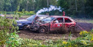 Dwa samochody rajdowe ścigają się na drodze pokrytej gałęziami. Czerwony samochód na zniszczoną maskę z przodu, widoczny jest silnik. Obok niebieski samochód, nad którym unosi się dym. W tle zielony las. 