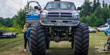 Samochód typu monster truck z wysokim zawieszeniem i bardzo dużymi kołami stoi na trawie. Mężczyzna oraz kobieta z dzieckiem na rękach oglądają samochód w środku. W tle zielone drzewa i pochmurne niebo.