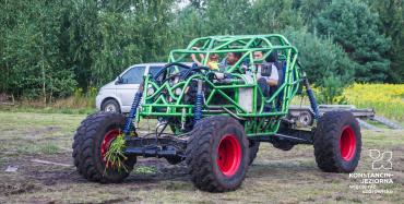 Trzy osoby, dwóch mężczyzn i dziewczynka jadą po trawie samochodem typu monster truck. Samochód zbudowany jest z zielonych metalowych rur połączonych ze sobą oraz z czterech dużych kół. W tle zielony las.