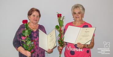 Dwie kobiety w średnim wieku stoją obok siebie. Każda w rękach trzyma dyplomy i czerwoną różę. W tle biała ściana.  