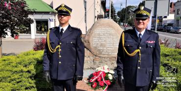 Dwóch strażników miejskich, ubranych w stroje galowe, stoi na baczność obok pomnika upamiętniającego bohaterów Bitwy Warszawskiej z 1920 roku. Pod pomnikiem zostały złożone biało czerwone kwiaty.