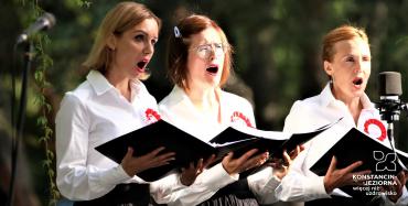Trzy kobiety – śpiewaczki, stoją obok siebie w rzędzie. Ubrane są w białe koszule. W rękach trzymają śpiewniki. 