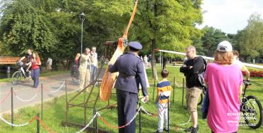 Park Zdrojowy. Na trawie stoi duże drewniane śmigło. Obok stoi mężczyzna ubrany w strój pilota, na głowie ma czapkę. Za nim stoją kobieta i mężczyzna oraz dwójka dzieci.