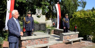 Cmentarz. Trzech mężczyzn w średnim wieku stoi przy pomniku ofiar drugiej wojny światowej. Jeden z mężczyzn ubrany jest w garnitur, pozostali mają na sobie mundury Straży Miejskiej. Po obu stronach pomnika na sztandarze wiszą biało-czerwone flagi. 