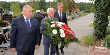 Cmentarz. Trzech mężczyzn w średnim wieku idzie obok siebie w rzędzie, ubrani są w garnitury. Jeden z mężczyzn trzyma w rękach wieniec z biało-czerwonymi kwiatami.
