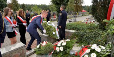 Cmentarz. Troje dzieci w wieku szkolnym – dwie dziewczynki i jeden chłopiec, składają kwiaty przed pomnikiem ofiar drugiej wojny światowej. Przy pomniku stoi mężczyzna ubrany w mundur Straży Miejskiej. Na pomniku leżą trzy wieńce z biało-czerwonymi kwiatami.