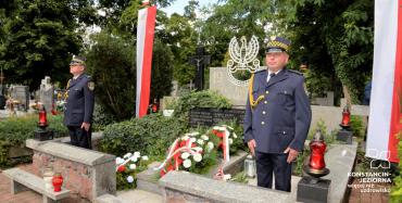 Cmentarz. Przy pomniku ofiar drugiej wojny światowej stoi dwóch mężczyzn w średnim wieku. Ubrani są w mundury Straży Miejskiej. Na pomniku leżą cztery wieńce z biało-czerwonymi kwiatami. Przed pomnikiem stoją cztery znicze, trzy czerwone i jeden biały. Po obu stronach pomnika na sztandarze wiszą biało-czerwone flagi. 