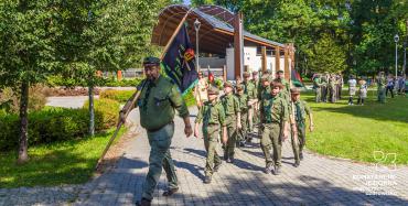 Park Zdrojowy. Mężczyzna ubrany w strój harcerza idzie po chodniku aleją. W ręku trzyma flagę z nazwą grupy harcerskiej. Za nim idzie grupa około dziesięciu osób – dzieci w wieku szkolnym i młodzież. Wszyscy są ubrani w harcerskie stroje. W tle amfiteatr i zielone drzewa.