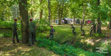 Park Zdrojowy. Grupa dziesięciu osób – dzieci w wieku szkolnym. Stoją obok siebie, rękach trzymają czarną linę, która jest przywiązana do dwóch drzew oddalonych od siebie. Wszyscy ubrani są w harcerskie stroje, na głowach mają bordowe berety.