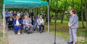 Po prawej stronie stoi elegancko ubrana kobieta w starszym wieku, przemawiająca do mikrofonu umieszczonego na stojącym przed nią statywie, na środku i lewej stronie pod namiotem z zielonym dachem na krzesłach siedzą elegancko ubrane osoby w różnym wieku, kilkanaście osób stoi także za namiotem, na otoczce dachu biały napis www.konstancinjeziorna.pl , wszyscy na alei parkowej, wokół zielone drzewa.