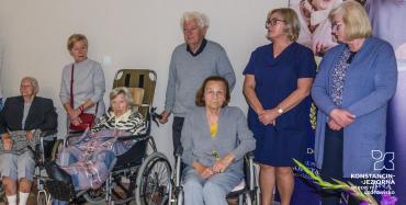 Sala. Trzy kobiety – seniorki siedzą na wózkach inwalidzkich. Obok stoją cztery osoby trzy kobiety w średnim wieku oraz mężczyzna – senior.