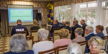 Sala. Około dwadzieścia osób siedzi na krzesłach. Z tyłu siedzą seniorzy. Z przodu siedzi kobieta oraz sześciu mężczyzn w średnim wieku. Jeden z nich ma kurtkę z napisem Policja.  Naprzeciwko nich jest projektor na którym jest wyświetlana prezentacja o Domu Seniora.