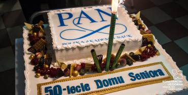 Sala. Na stoliku stoi duży biały tort z napisem Polska Akademia Nauk oraz 50-lecie Domu Seniora. Tort ozdobiony jest dookoła owocami oraz czekoladowymi ozdobami.