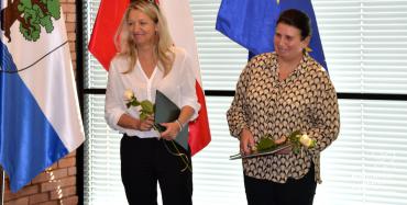 Sala konferencyjna. Dwie kobiety w średnim wieku stoją obok siebie, w rękach trzymają dyplomy i białe róże. W tle trzy flagi: flaga Konstancina-Jeziorny, flaga Polski i flaga Unii Europejskiej oraz okno z żaluzjami.