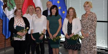 Sala konferencyjna. Siedem osób stoi obok siebie w rzędzie – sześć kobiet i jeden mężczyzna. Cztery kobiety w rękach trzymają dyplomy i białe róże. W tle trzy flagi: flaga Konstancina-Jeziorny, flaga Polski i flaga Unii Europejskiej.