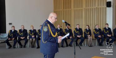 Sala konferencyjna. Mężczyzna w średnim wieku, stoi przy mikrofonie, w rękach trzyma kartkę. Ubrany jest w granatowy mundur Straży Miejskiej, na nosie ma okulary. Za nim siedzi dziesięć osób na krzesłach. Wszyscy ubrani są w granatowe mundury Straży Miejskiej.