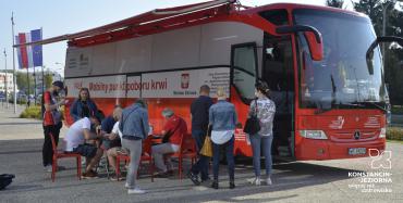 Miasto. Na chodniku stoi czerwony autokar, z boku ma duży napis Mobilny punkt poboru krwi, obok jest godło Polski i napis Minister Zdrowia.  Przed autokarem stoi sześć osób oraz na krzesłach przy stoliku siedzą cztery osoby.