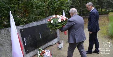 Park. Przed pomnikiem ofiar katyńskich stoi dwóch mężczyzn w średnim wieku. Jeden z nich trzyma wiązankę z białymi kwiatami. Przy pomniku stoją znicze i leżą biało-czerwone kwiaty. Po obu stronach pomnika stoją na stojaku biało-czerwone flagi.